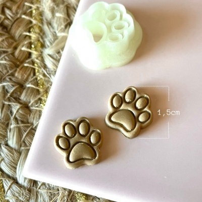 Brincos dourados em forma de pata de animal com molde branco sobre superfície rosa clara e fundo de fibra natural trançada