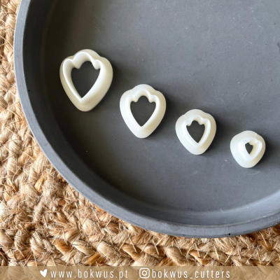 Quatro cortadores de biscoitos em formato de coração, brancos, dentro de um prato preto sobre base de têxtil trançada