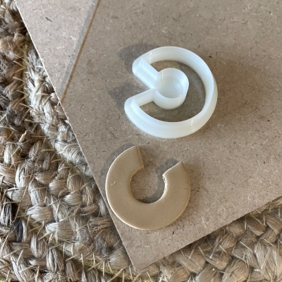 Dois anéis em forma de C, um branco e outro dourado fosco, sobre superfície castanha e tecido trançado