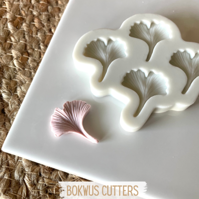 Molde de silicone branco com cavidades em forma de folha de ginkgo e peça rosa claro sobre superfície branca