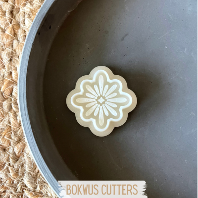 Cortante em forma de flor bege clara com padrões, sobre prato cinzento e fundo têxtil bege