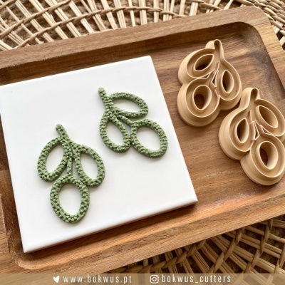 Cortadores de biscoito em forma de ramo ao lado de biscoitos verdes no tabuleiro, sobre textura de rattan.