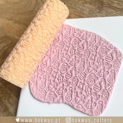 Rolo de massa cor laranja clara com relevo de figuras natalícias e massa cor rosa com relevo de Natal em superfície branca e madeira