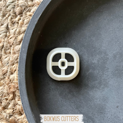 Cortador de massa quadrado branco com cruz numa base preta sobre esteira natural.