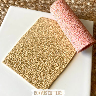 Rolo texturizador rosa com padrão floral sobre massa bege em superfície branca