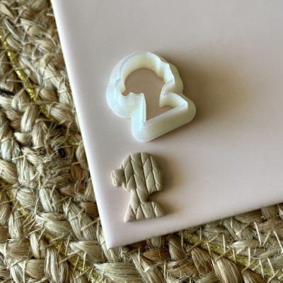 Cortador branco em forma de boneca e peça bege texturizada sobre superfície rosa e tapete de fibras naturais