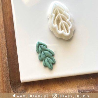 Cortador de massa em formato de folha branco e peça de massa azul com manchas pretas num prato branco sobre uma superfície de madeira.