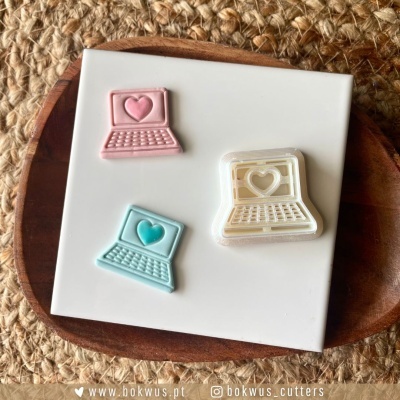 Três cortadores de biscoitos em forma de portátil em rosa, azul e branco sobre placa branca e bandeja de madeira