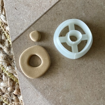 Peças pequenas, uma dourada em forma irregular e outra branca plástica redonda com orifício, sobre superfície de madeira.