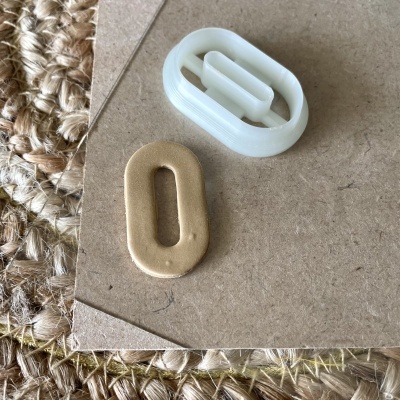 Duas pegas ovaladas, uma dourada em metal e outra branca em plástico, sobre cartão castanho e tapete trançado