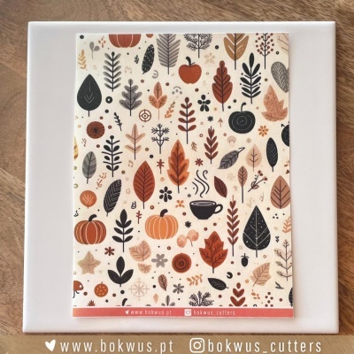 Cartão decorativo com padrão de folhas e abóboras em cores outonais sobre placa branca numa mesa de madeira.