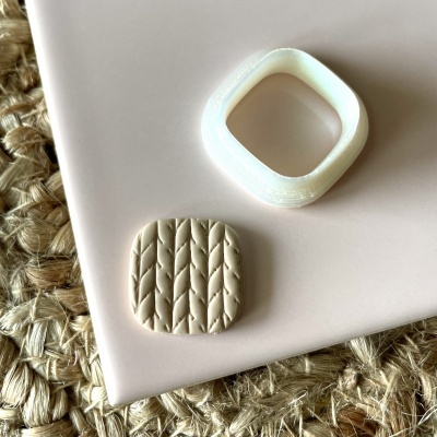 Duas peças quadradas, uma branca de plástico e outra creme com texturas, em fundo rosa claro e tapete fibras naturais