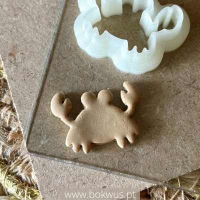 Cortador de biscoitos branco em forma de caranguejo com biscoito dourado no mesmo formato numa superfície de madeira clara