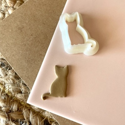 Cortante branco em forma de gato sentado e figura cortada em pasta dourada sobre fundo rosa claro