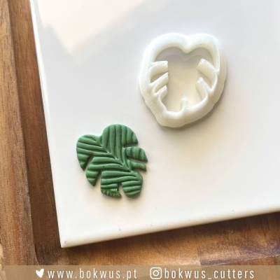 Cortador de massa em forma de folha com peça verde cortada ao lado sobre superfície branca e madeira.