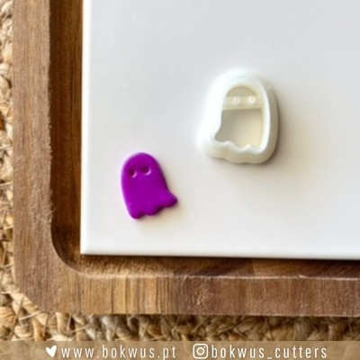 Cortadores de biscoitos em forma de fantasmas, um branco e outro roxo, sobre superfície branca e tabuleiro de madeira.