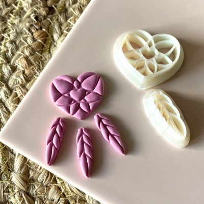 Moldes de plástico branco para cortar massa em forma de coração e folhas, com peças cor rosa claro, sobre superfície bege e tapete de juta ao lado.