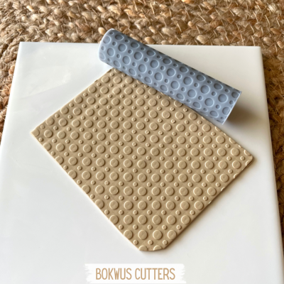 Rolo texturizador azul e folhas de argila bege com padrão circular sobre superfície branca e fundo têxtil