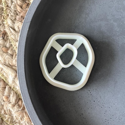 Objeto plástico branco geométrico sobre superfície escura e textura porosa, com tapete de fibras naturais ao lado