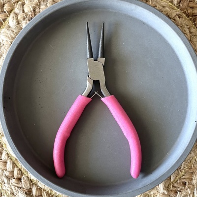 Alicate de pontas finas com punhos cor de rosa em travessa cinzenta redonda sobre superfície bege de tecido