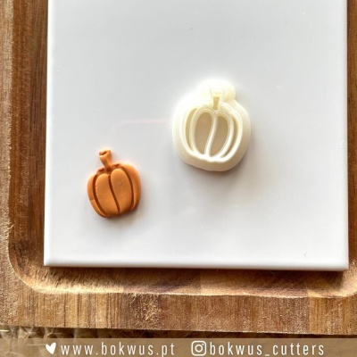 Dois cortadores de biscoito em forma de abóbora, um laranja e outro creme, sobre azulejo branco e tábua de madeira