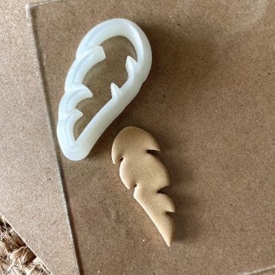 Cortador plástico branco e peça de couro bege em forma de pena sobre cartão marrom