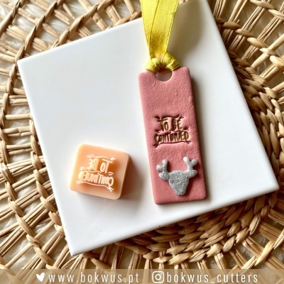 Cubo e placa rosa de argila com texto dourado e lacre prateado sobre prato branco e cesto trançado