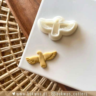 Cortador e bolacha em forma de pássaro em voo sobre superfície branca com fundo de vime