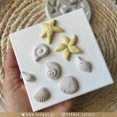 Estrelas do mar e conchas em material branco e amarelo sobre base branca, mão segurando, superfície de corda trançada ao fundo.