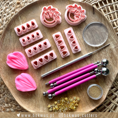 kit de cortadores e ferramentas de modelagem rosa e prateadas com moldes e pequenas pérolas douradas num prato de madeira sobre superfície trançada