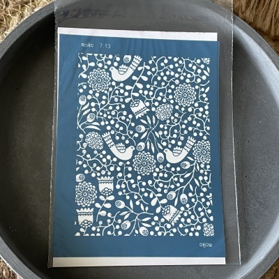 Stencil de plástico azul com padrão branco de aves e flores dentro de saco plástico sobre prato metálico cinzento