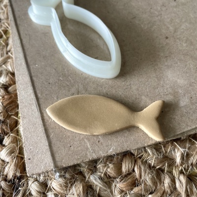 Cortador de massa branco em forma de peixe com massa bege cortada similar em superfície de cartão e tapete de fibras naturais.
