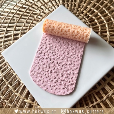 Rolo texturizador rosa coral com padrão floral sobre superfície branca e fundo de cesto natural