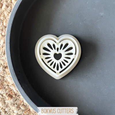 Cortador de biscoito em forma de coração branco com padrão recortado sobre prato preto.