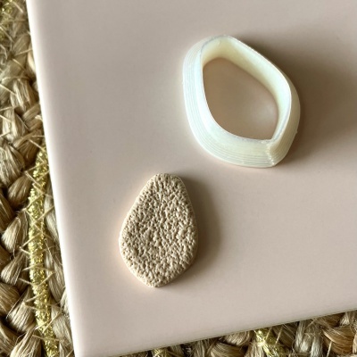 Dois objetos em formato de gota, um branco translúcido e outro bege texturizado, sobre superfície rosa clara com fundo de palha