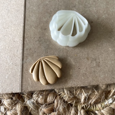 Dois objetos em forma de concha, um branco e outro de madeira, sobre superfície de cartão castanho e fibra natural.