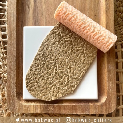 Rolo texturizador e argila com padrão de folhas em placa branca sobre tábua de madeira