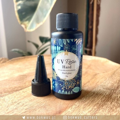 Garrafa azul escuro de resina UV Hard com rótulo decorado e texto, sobre mesa de madeira, tampa preta ao lado.