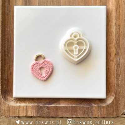 Molde de cortar bolachas e bolacha em forma de cadeado com coração, em plástico branco e rosa, sobre azulejo branco e tabuleiro de madeira