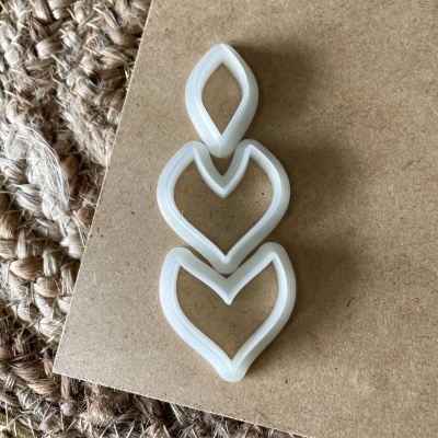 Três cortadores de biscoitos brancos em formato de folhas/corações sobre papelão e tapete de fibras naturais.