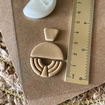 Brincos bege geométricos sobre cartão castanho ao lado de régua amarela em fundo de tecido grosso