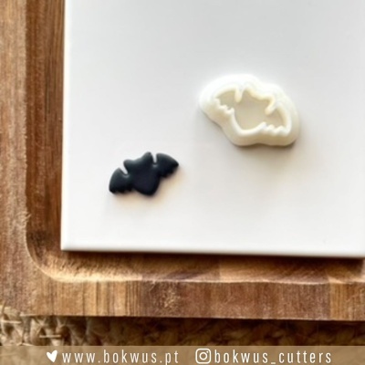 Formas de corte para bolachas em forma de morcego, uma preta e uma branca, sobre superfície branca e tabuleiro de madeira.