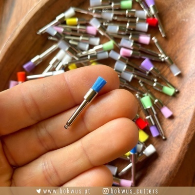 Broca de escova pequena azul com haste metálica segurada à frente, várias brocas coloridas dentro de tigela de madeira ao fundo.