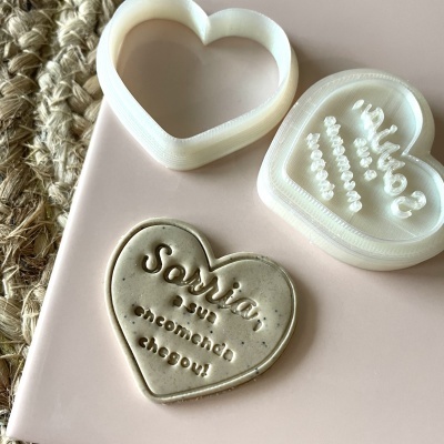 Cortantes de coração em plástico branco e biscoito com frase gravada sobre superfície rosa