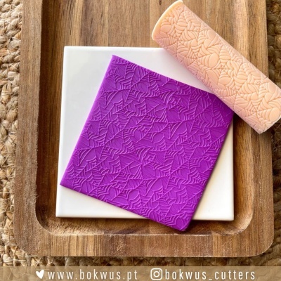 Placa texturizada roxa e rolo de massa com padrão de folhas sobre tabuleiro de madeira