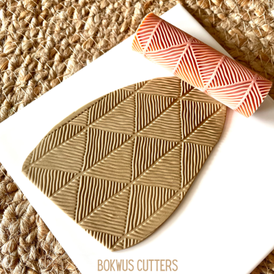 Rolo texturizador com padrão de triângulos e placa de argila texturizada sobre superfície branca e tapete de fibras.