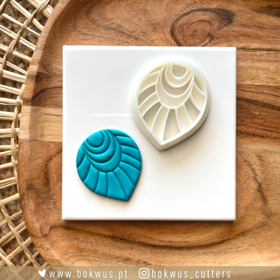 Cortador branco de pasta de polímero e peça azul com padrão de folha sobre uma placa branca e fundo de madeira com tapete de palha.
