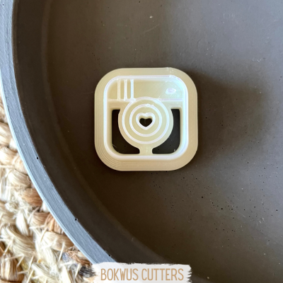 Cortador branco em forma de câmara fotográfica com coração no centro sobre superfície cinza.