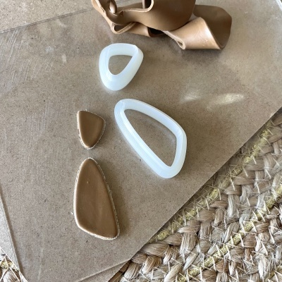 Brincos assimétricos bege e branco sobre superfície castanha textil com tira de couro.