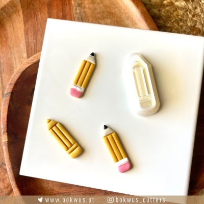 Três lápis decorativos amarelos e um cortador de plástico branco em forma de lápis sobre placa branca e base de madeira.